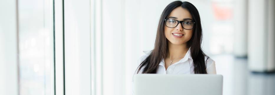Confident woman with long dark hair and glasses holding laptop near bright windows in a modern office environment.
