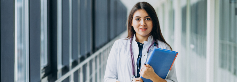Young woman in white coat holding blue folders in bright hallway, looking confident in a medical or academic setting.